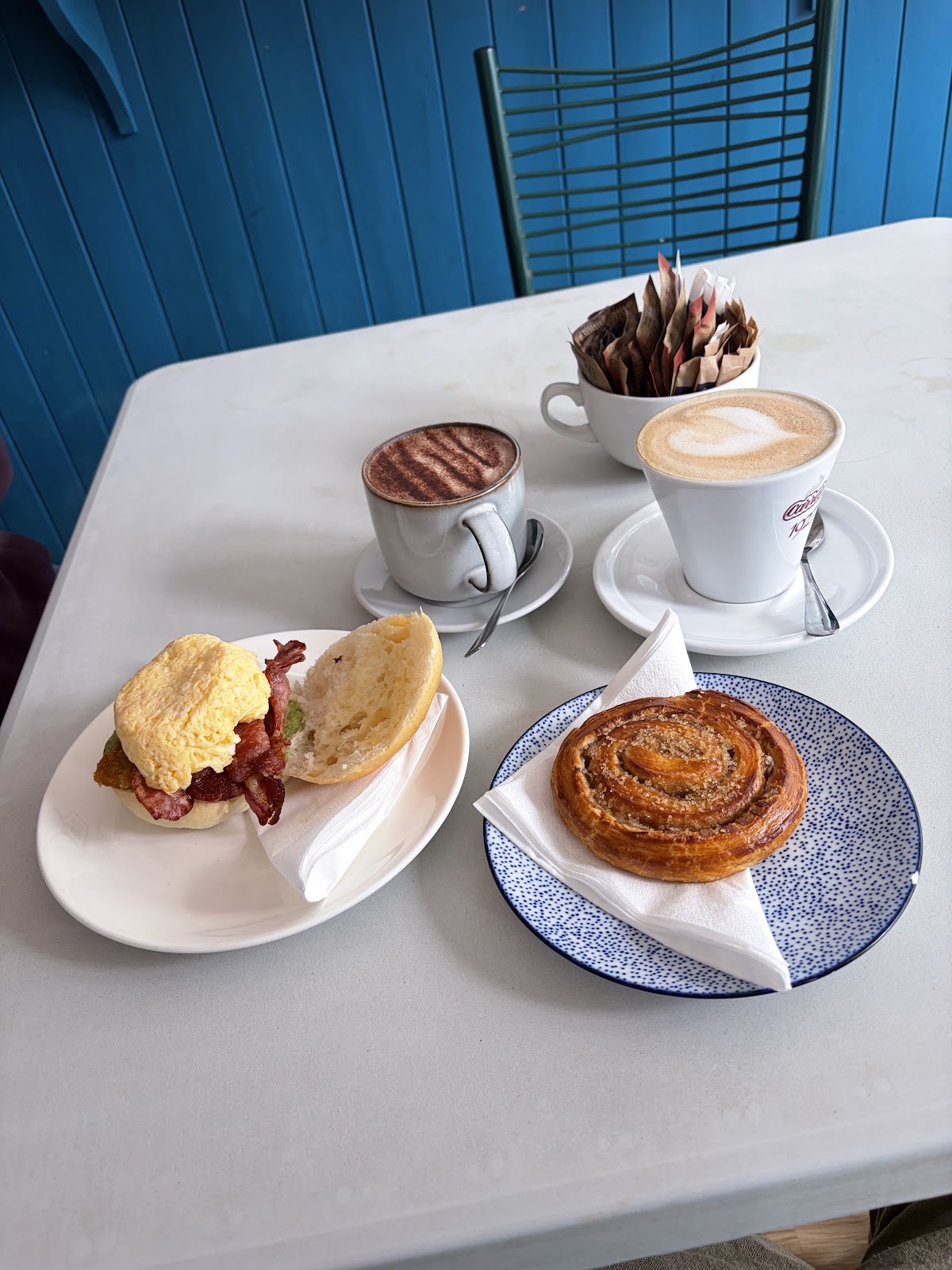 A roll with scrambled egg, bacon and hash brown next to a sugar-crusted cinnamon swirl on a blue spotted plate, a latte and a mocha, against a blue tongue-and-groove wall.