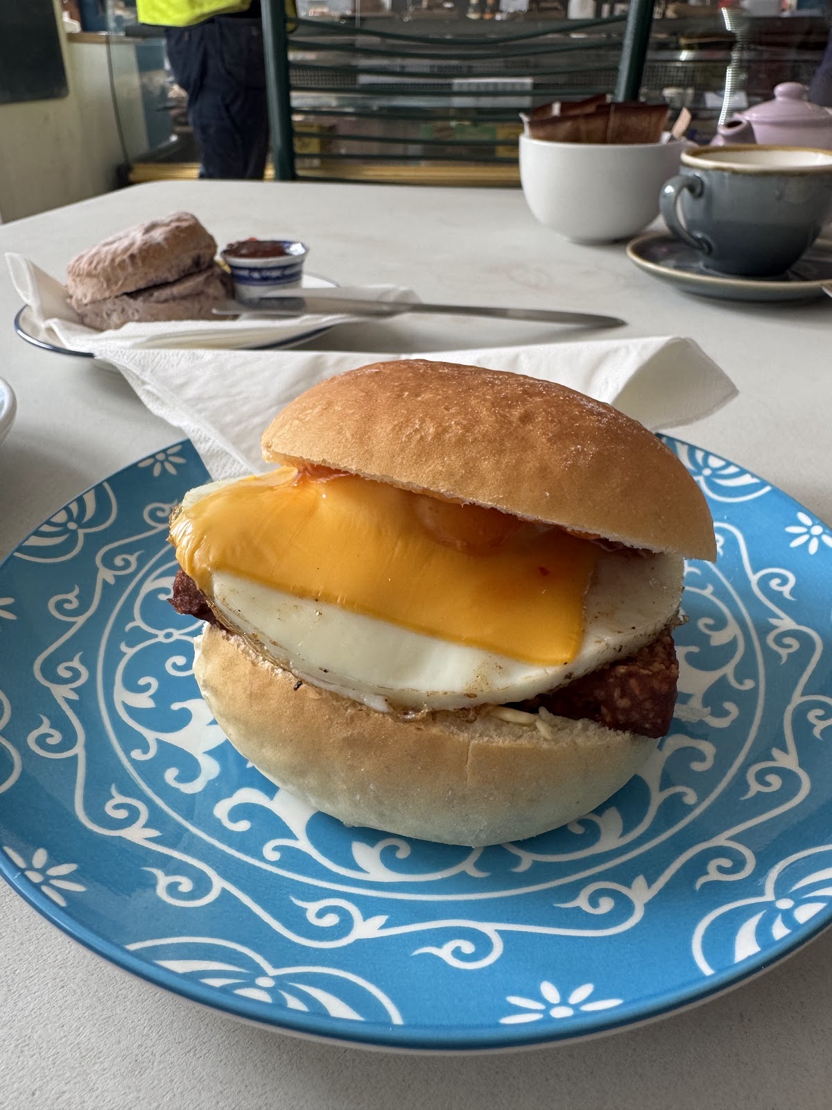 A soft white roll with fried egg, cheese and square sausage on a blue and white patterned plate, with a scone and jam behind.